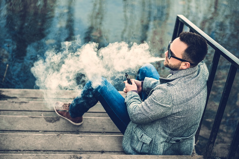 Man sitting by water smoking Cannabis vape cartridge