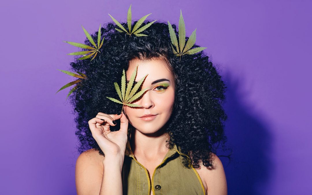 Girl with Cannabis Leaves in her hair, holding a Cannabis leaf up to her eye.