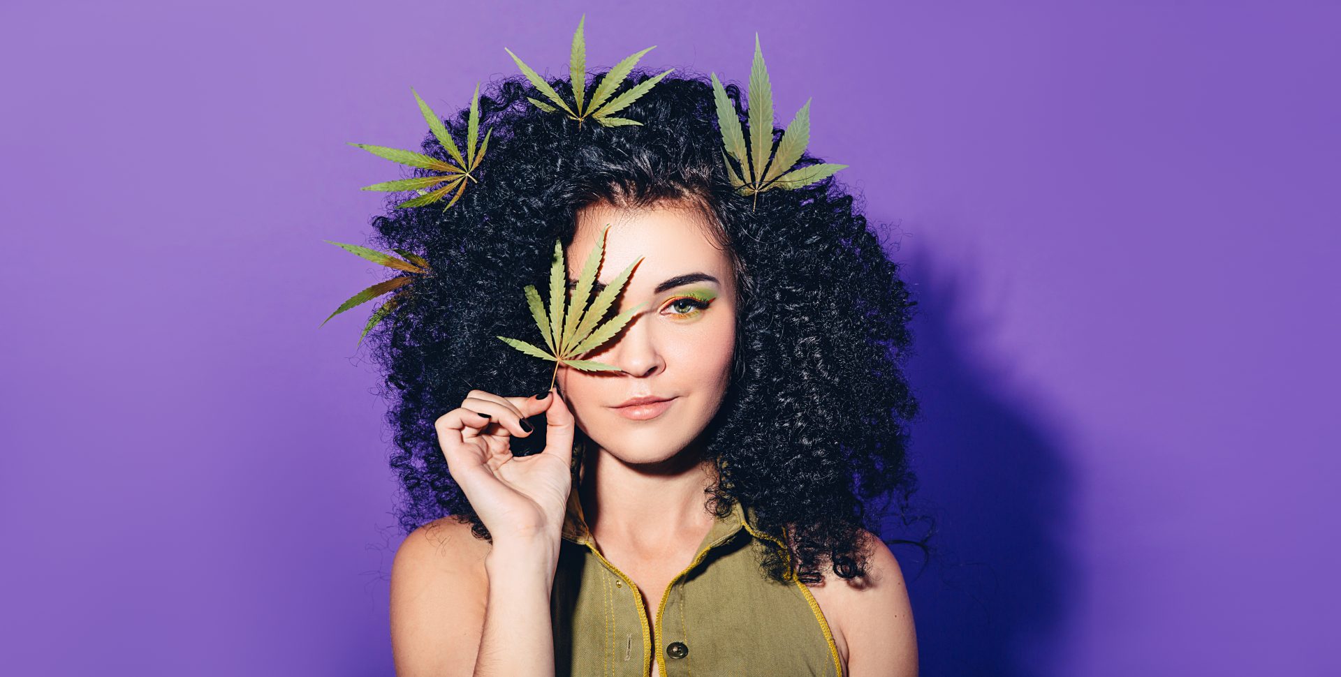 Girl with Cannabis Leaves in her hair, holding a Cannabis leaf up to her eye.
